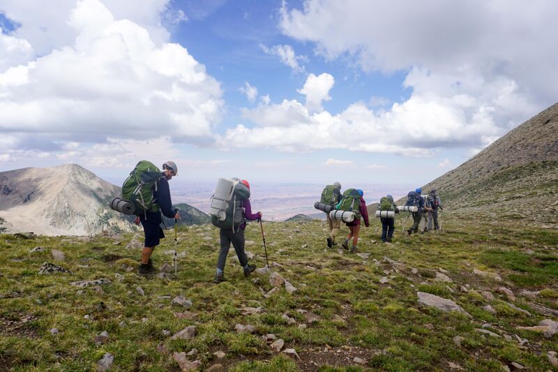 A group of hikers is trekking across a grassy, mountainous landscape under a partly cloudy sky. They are equipped with large backpacks, suggesting a multi-day trip. The terrain appears rugged, with a mix of green vegetation and rocky patches. In the background, a mountain peak is visible, adding to the scenic backdrop of their outdoor adventure.